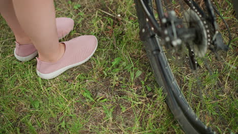 close-up of bicycle wheel and pedal with person wearing pink sneaker removing stand, set in lush green park with blurred background of grass and trees