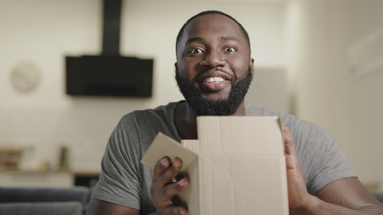 Happy black man sitting on couch with open box. Surprised young guy holding box.