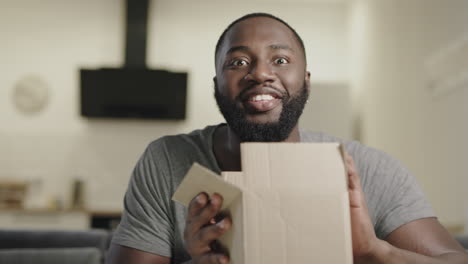 Happy-black-man-sitting-on-couch-with-open-box.-Surprised-young-guy-holding-box.
