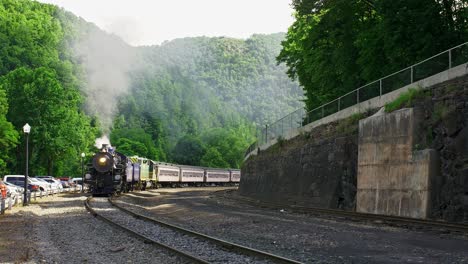 a steam train chugs along a picturesque mountain route while releasing plumes of steam, surrounded by lush greenery and parked cars. the sun shines brightly above, enhancing the vibrant scenery.