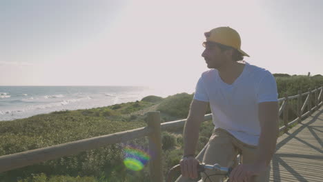 Männlicher-Radfahrer-Auf-Einer-Promenade-In-Richtung-Strand-Mit-Blick-Auf-Das-Meer