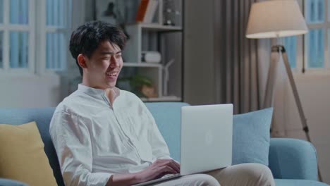 Smiling Asian Man Having Video Call On Laptop Computer While Sitting On Sofa In The Living Room