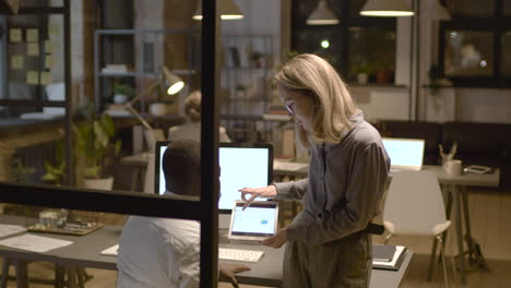 rear view of american man sitting at desk while talking with female coworker who is showing him graphics on the tablet device in the office