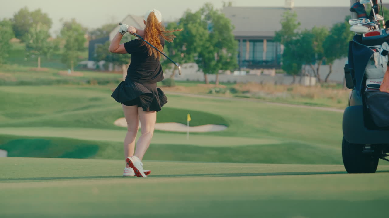 Female golfer hits an iron approach shot off the fairway next to a golf cart with the flagstick visible in the background. A focused moment showcasing aim and precision heading toward the green.