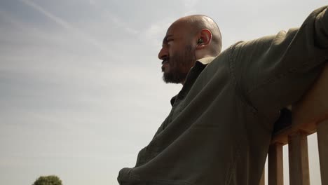 bald indian male sat on bench listening to music on earphones looking side to side pondering in thought