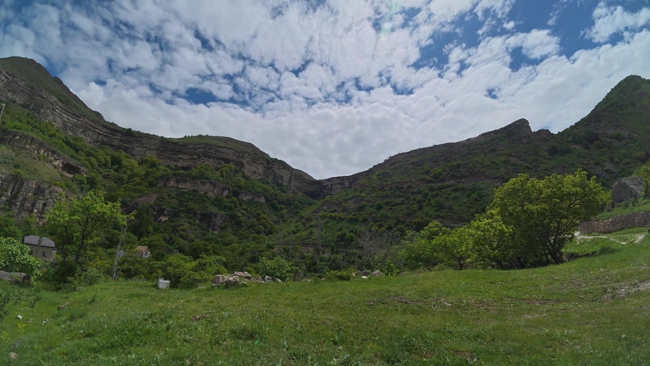 Un time lapse cattura nuvole bianche che si muovono attraverso un cielo blu sopra una lussureggiante valle di montagna verde con pendii ripidi e rocciosi e alberi sparsi.