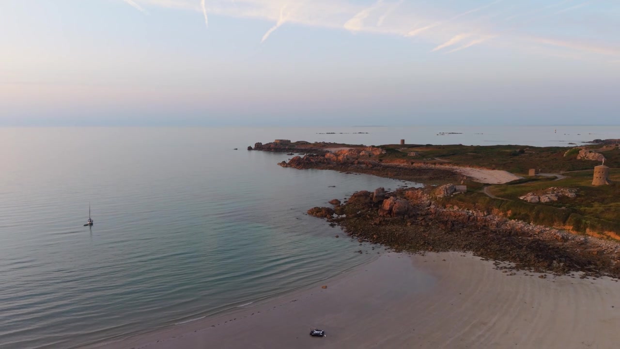 Guernsey, Isole del Canale. Filmato dell'ora d'oro che rivela una spiaggia di sabbia dorata con mare calmo e limpido e onde dolci lungo una costa rocciosa frastagliata, con il cielo che diventa rosa e un orizzonte nebbioso.