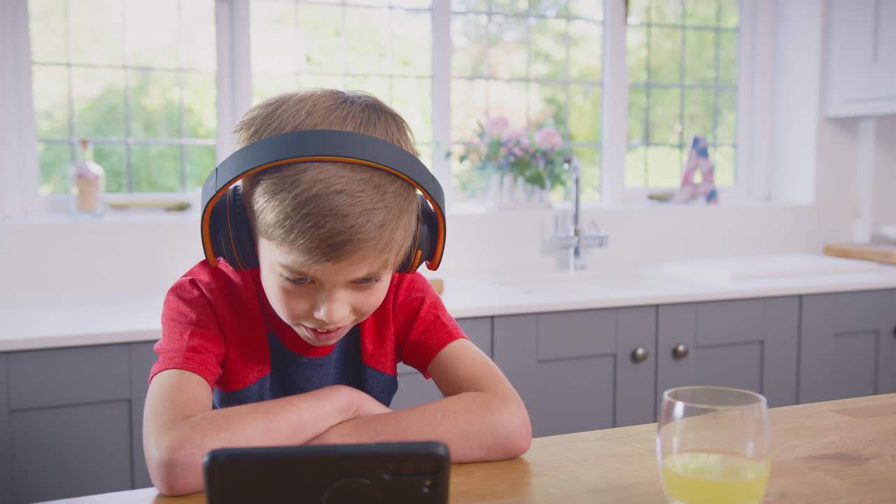 niño en casa en la cocina viendo una película o un programa en el teléfono móvil usando auriculares inalámbricos