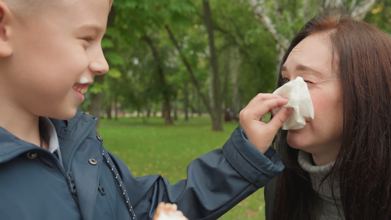 Ragazzo asciuga il viso di una donna con un fazzoletto su una panchina del parco, offrendo un cono gelato e un sorriso gentile, un gesto confortante e una tranquilla preparazione per la routine del rientro a scuola in un ambiente esterno verdeggiante.