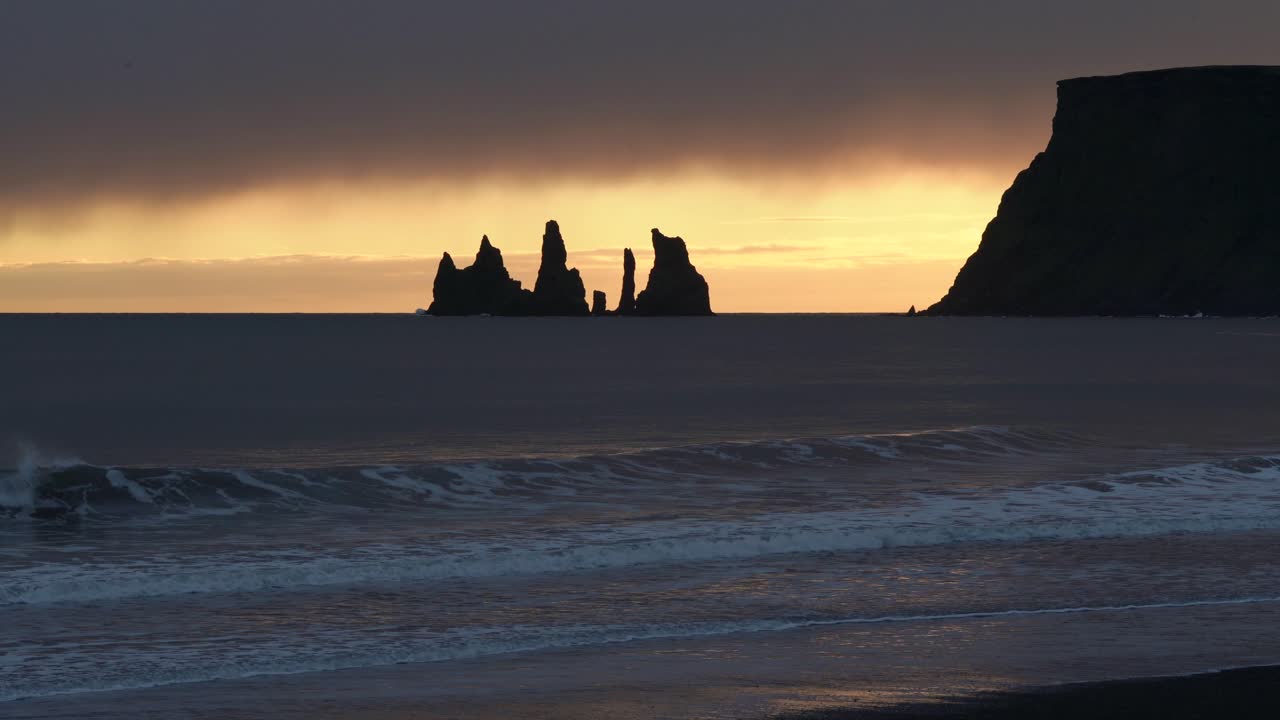 amplia silueta de reynisdrangar pilas de mar de basalto al lado de la montaña durante la luz del atardecer en islandia - olas que llegan a la playa durante el cielo misterioso - formación rocosa en el fondo