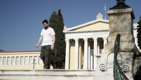 bearded man walks in front of athens historic zappeion sunny day in white shirt