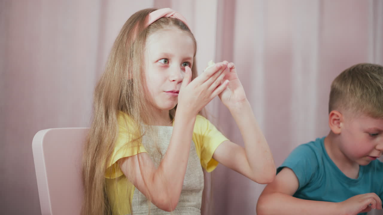 Smiling girl in yellow dress playfully holding and showing dough creation while her young brother seated beside her remains focused on shaping dough during fun baking at home with soft background