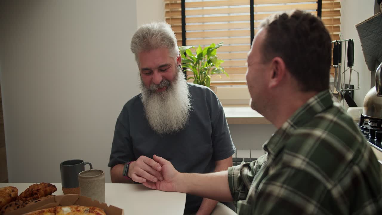 Over the shoulder A happy elderly man with gray hair with a lush gray beard strokes the hand of his brunette boyfriend in a checkered green shirt while sitting in the kitchen at the table and cheering during his communication