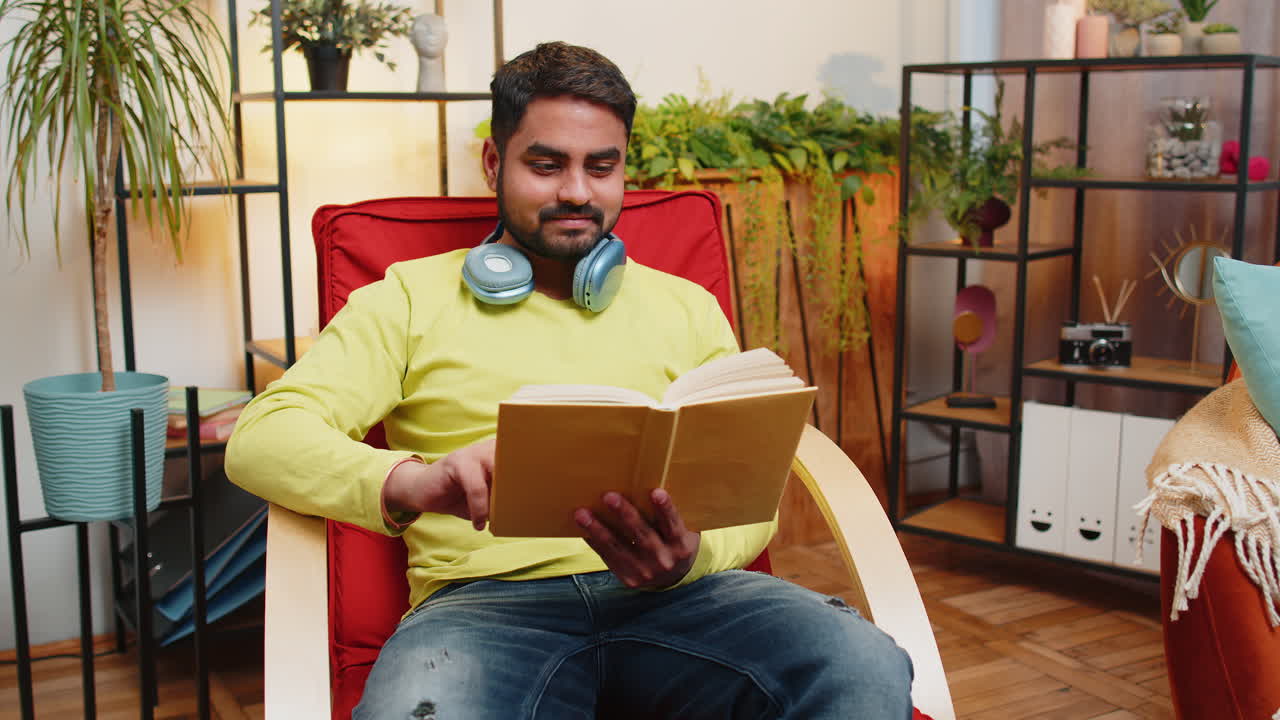 Indian man reading interesting book turning pages smiling enjoying literature taking a rest on chair
