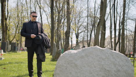 sad man in black suit walking in a graveyard, taking sunglasses off and looking at a tombstone sorrowfully