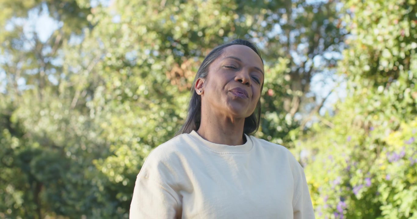 Mujer afroamericana inclinándose hacia atrás sintiendo el sol, bajando la barbilla y sonriendo en un jardín con una blusa color crema.