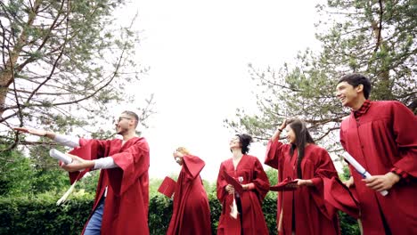 low shot of happy graduates throwing mortarboards in air, laughing and celebrating graduation on college campus. education, success and modern youth concept.