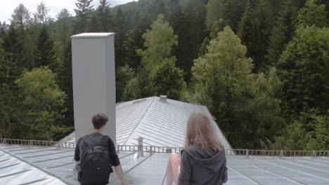 two people sitting on the roof of a big building surrounded by trees in slow-motion