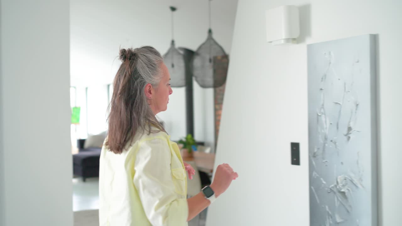 Woman is carrying large canvas toward white wall, lifting and aligning it near switch to center