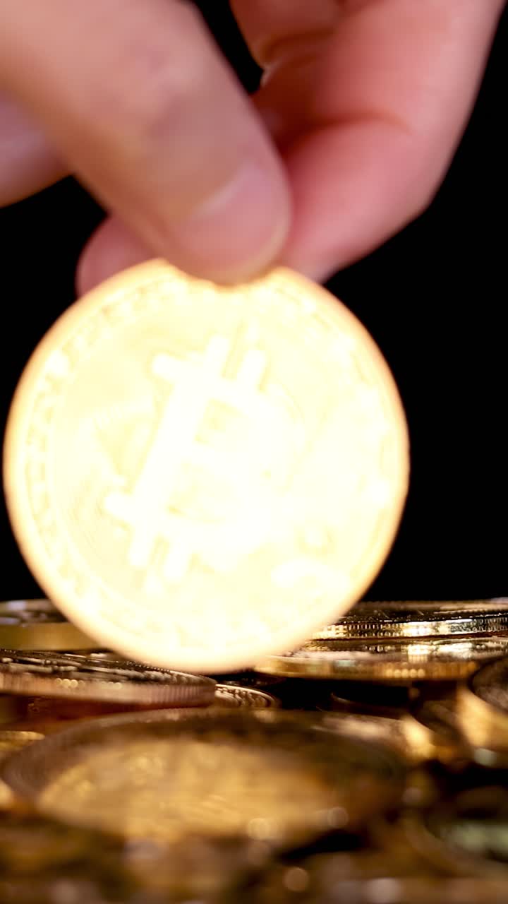 手が積み重ねられたトークンの上で輝く金色のビットコインをひっくり返す。ドラマチックな照明、クローズアップ、黒い背景。