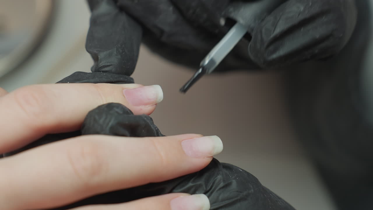 Close-up of nail technician wearing black glove precisely applying clear hardener on client fingernail using applicator brush, showcasing careful nail treatment procedure in salon environment