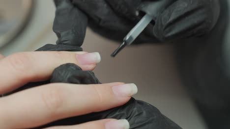 close-up of nail technician wearing black glove precisely applying clear hardener on client fingernail using applicator brush, showcasing careful nail treatment procedure in salon environment
