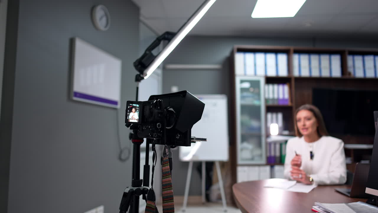 Professional camera and light set on tripod in front of the desk where speaker sits. Female blogger records a video for blog.