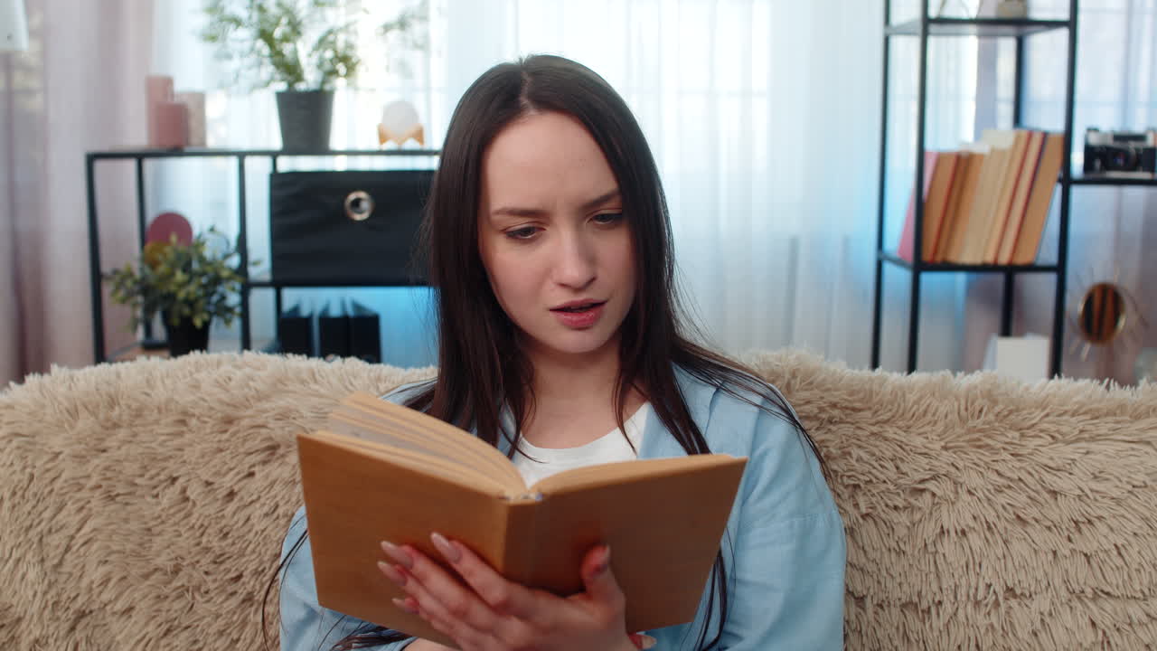 Young excited woman reading book on sofa deeply immersed emotionally reacting changing expressions