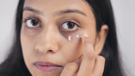 close up shot of an indian young lady applying moisturizing cream under eyes in white background, skin care concept.