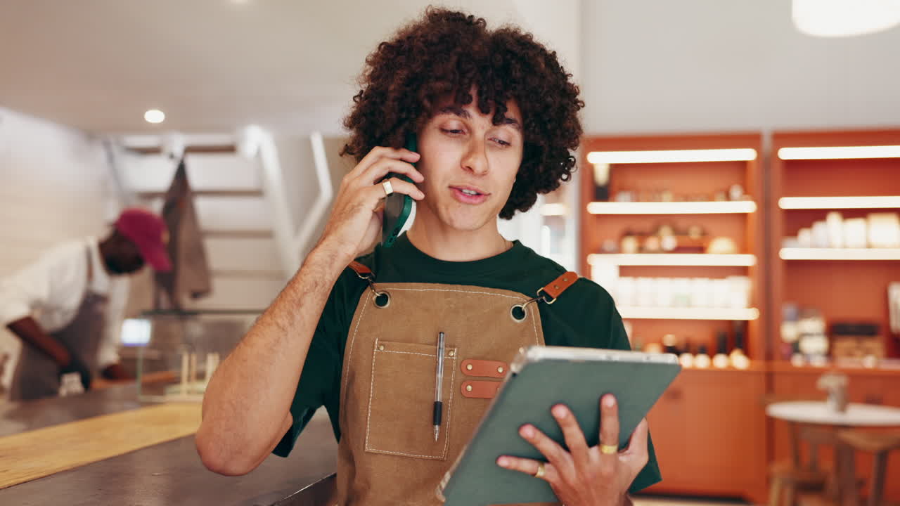 Man in apron talking on phone while holding tablet in coffee shop
