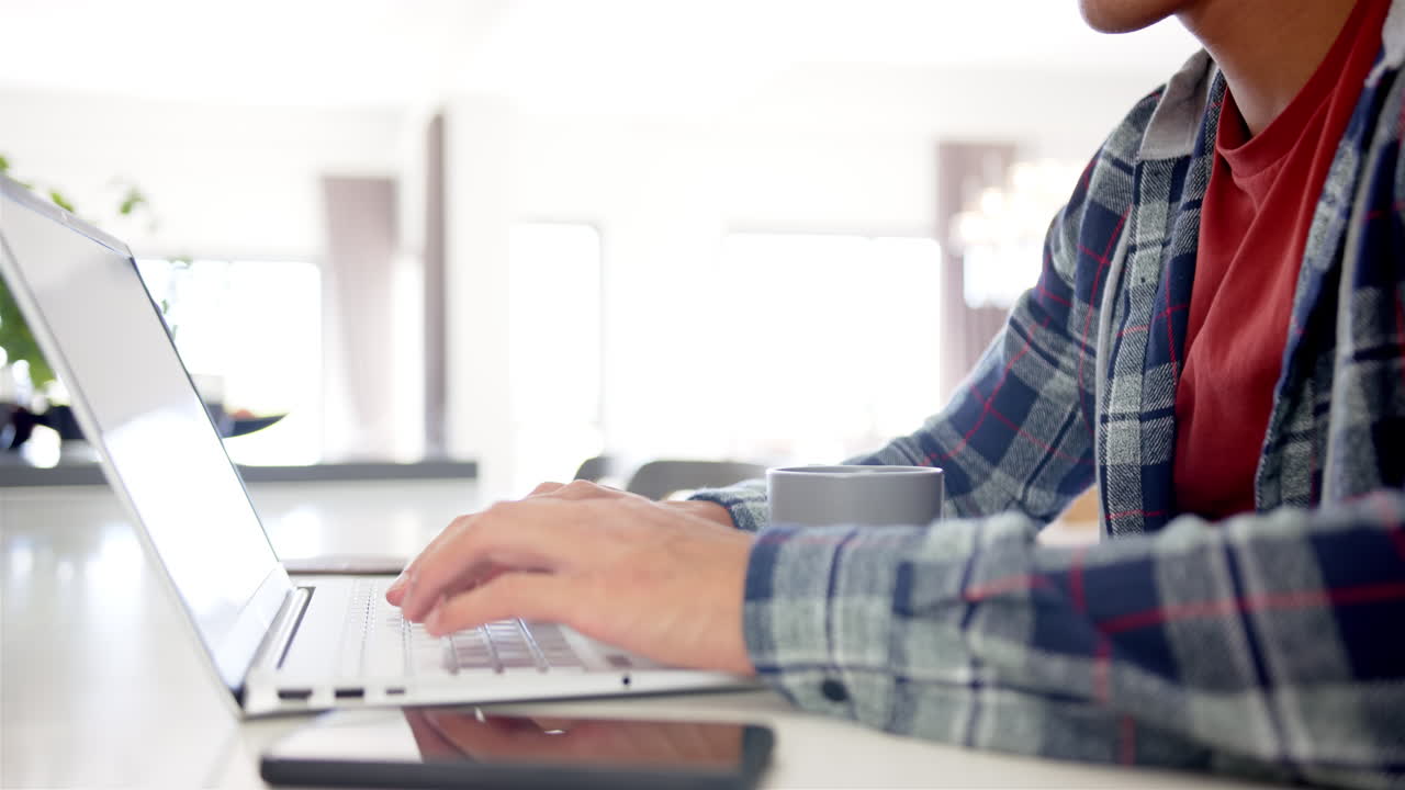 Typing on laptop, man working from home with smartphone and coffee mug nearby