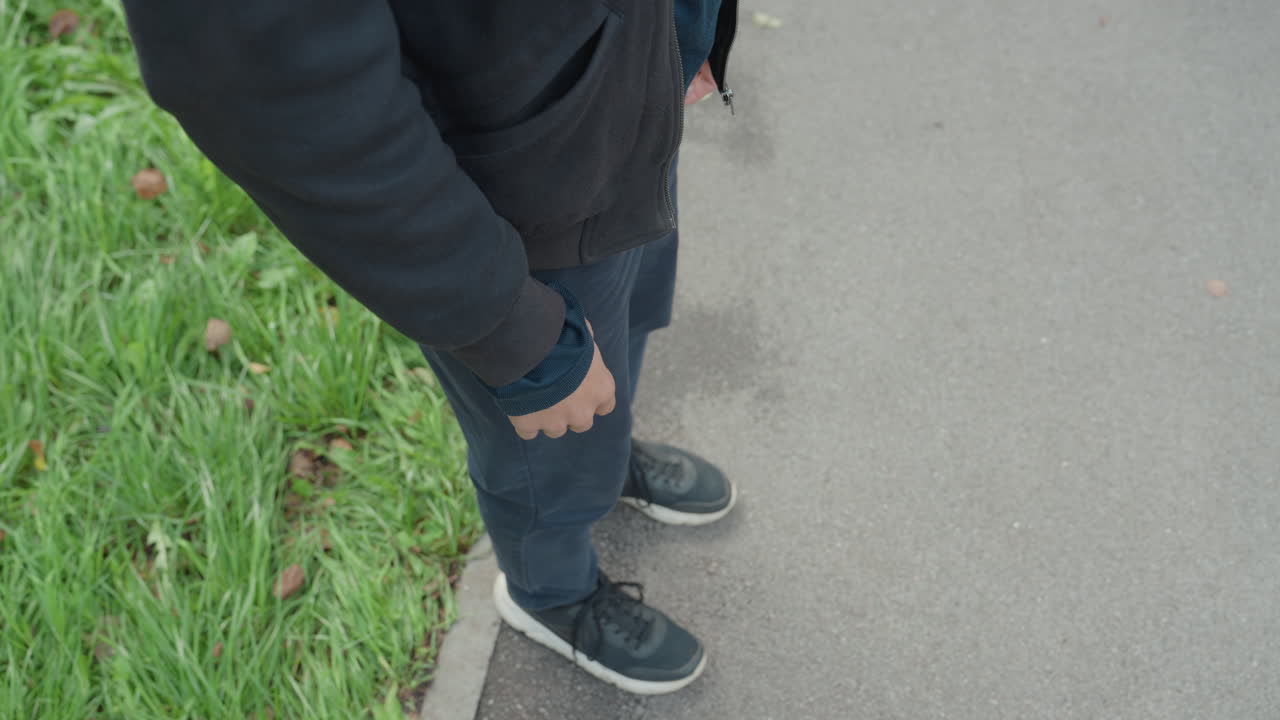 seorang pria berdiri di trotoar dekat rumput, tangan di saku, jaket dan sepatu kets berwarna gelap, tampilan taman kota dari atas ke bawah, perubahan berat badan yang halus, daun musim gugur di jalan setapak, suasana sinematik yang melukiskan kesendirian
