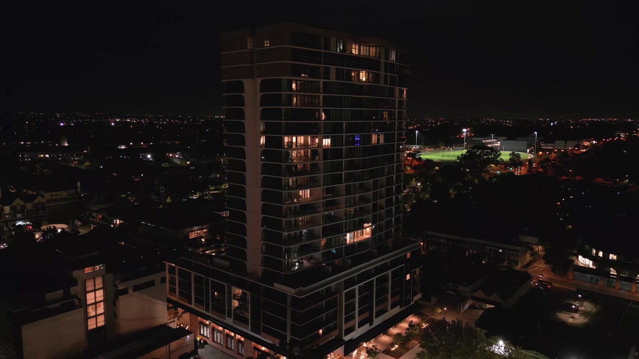Ripresa orbitale attorno a una moderna torre residenziale illuminata di notte, con appartamenti luminosi e luci della città, e un ovale sportivo illuminato a giorno che si estende attraverso il sobborgo buio. Subiaco, Perth, Australia