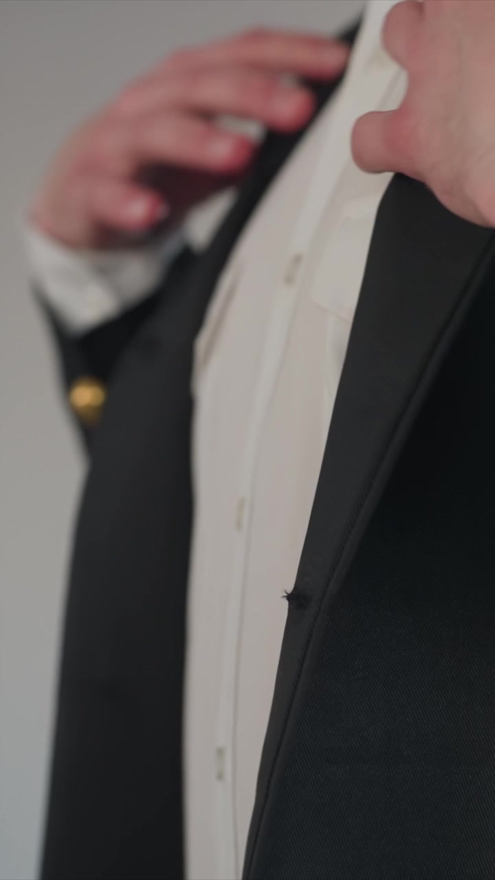 Primo piano di un uomo che sistema la sua camicia bianca e il suo abito nero. I dettagli includono polsini, spille da bavero e bottoni dorati. Lo sfondo semplice enfatizza le regolazioni dell'abbigliamento formale.