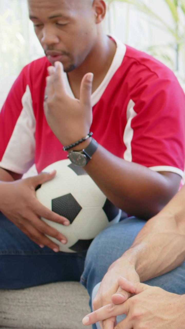 Vídeo vertical: Homem de camisa de futebol vermelha reagindo a um gesto, segurando uma bola de futebol enquanto conversa em casa.