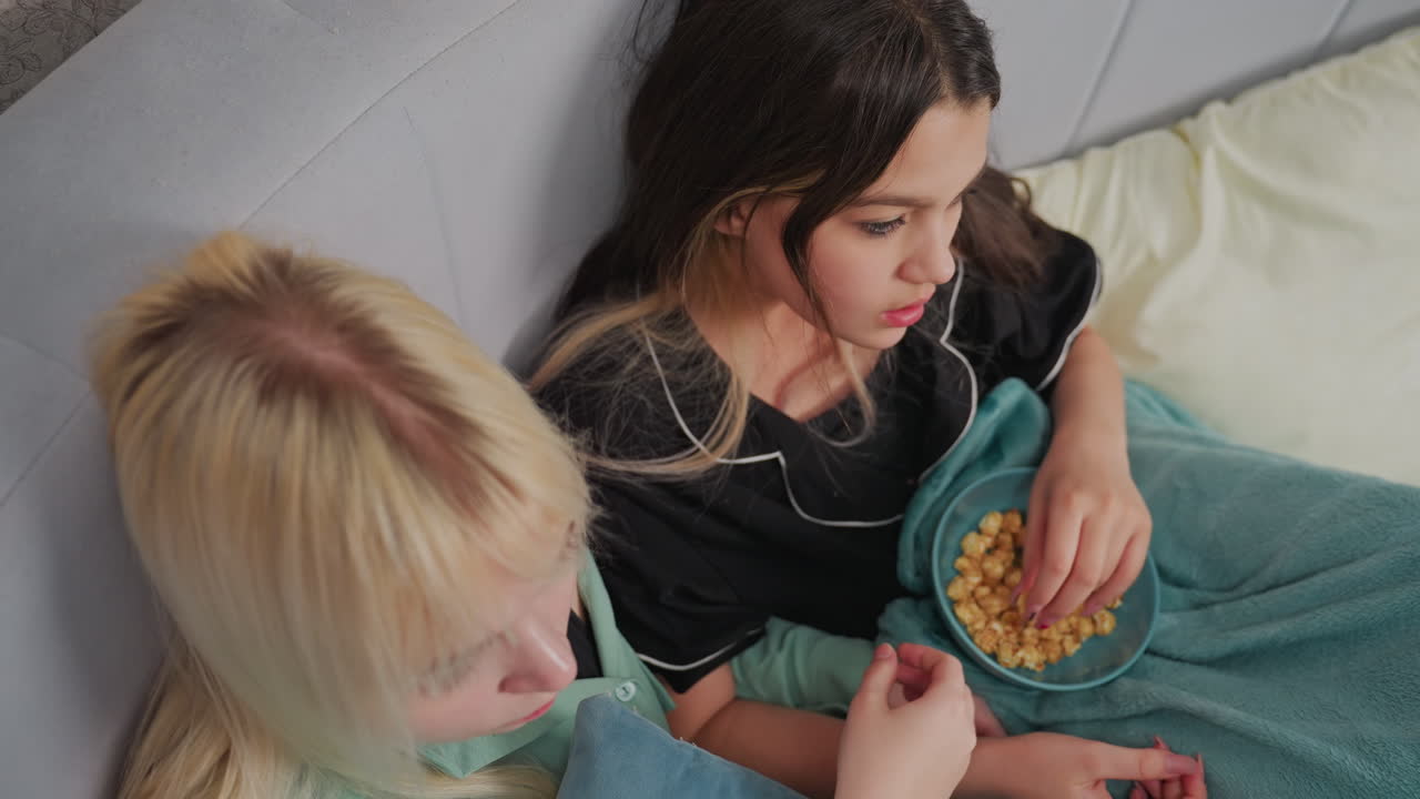 Top down view of two young girls sitting side by side on bed while eating popcorn from bowl, one girl feeds herself while other prepares to take popcorn