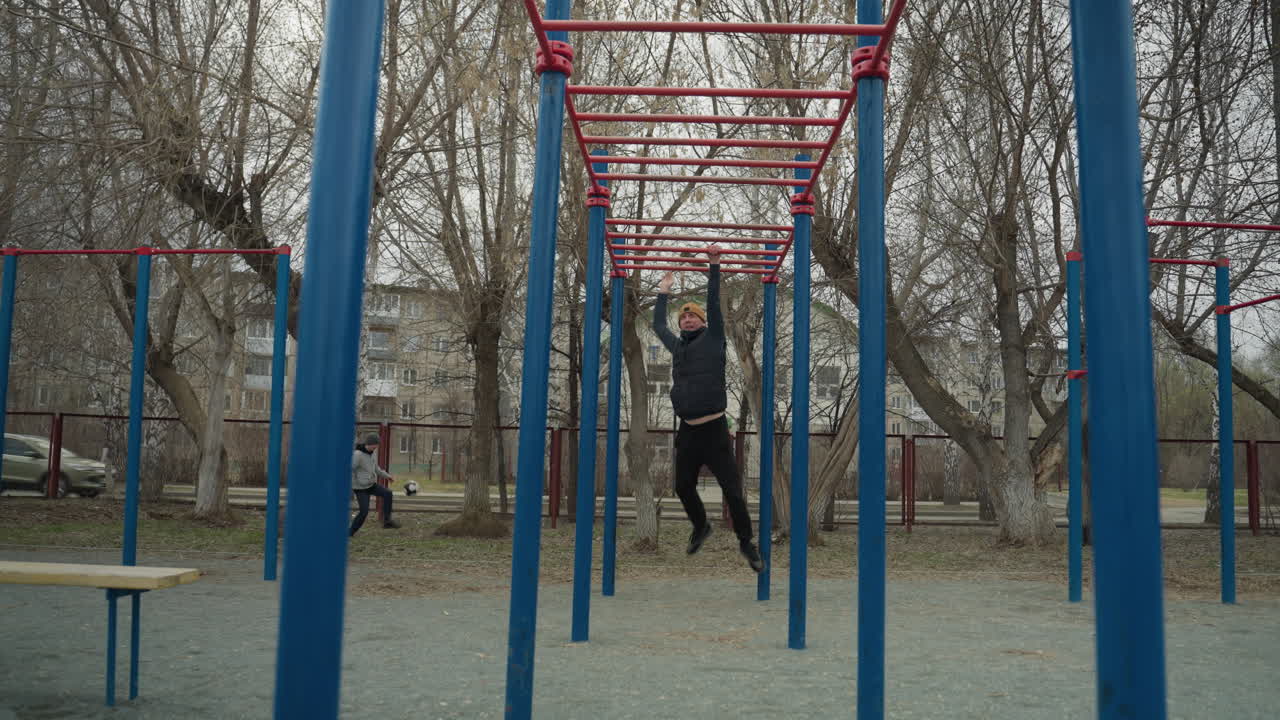 A coach jumps up and grabs a red iron bar, moving along it as he swings, in the background, a boy plays football, and cars pass by, with residential buildings and bare trees visible across the road