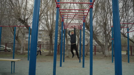 a coach jumps up and grabs a red iron bar, moving along it as he swings, in the background, a boy plays football, and cars pass by, with residential buildings and bare trees visible across the road