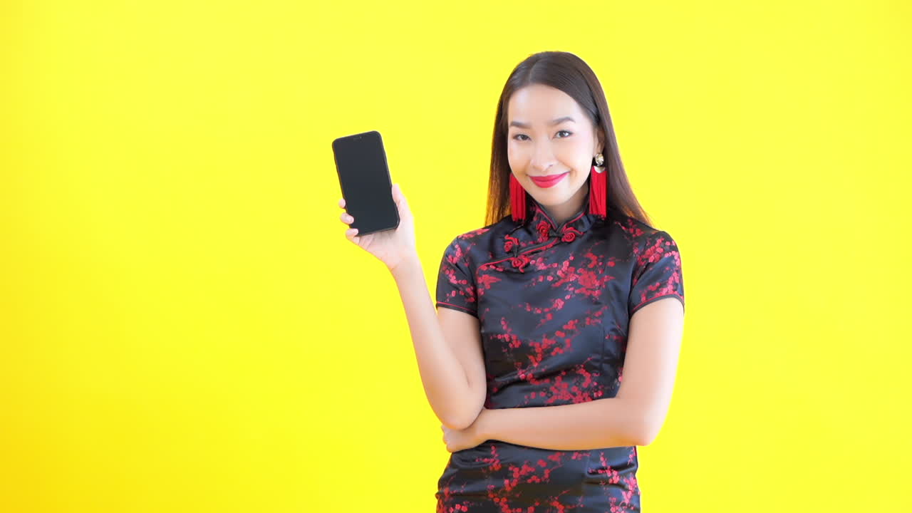 An attractive Asian woman standing with her arms crosses raises a smartphone and smiles. Copy space
