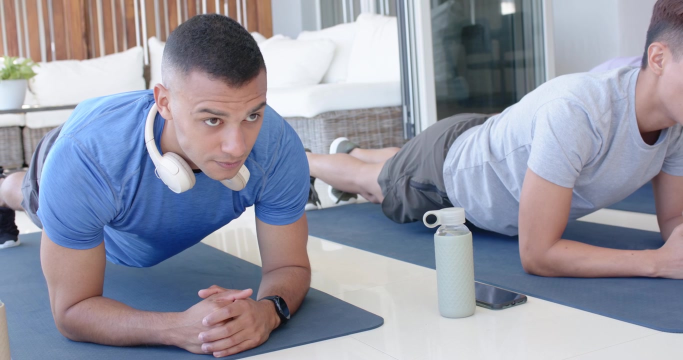 Diverse male friends after warmup, holding forearm planks at home with water bottle, phone for core