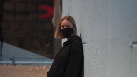 young lonely female with facemask behind wall in urban exterior, slow motion