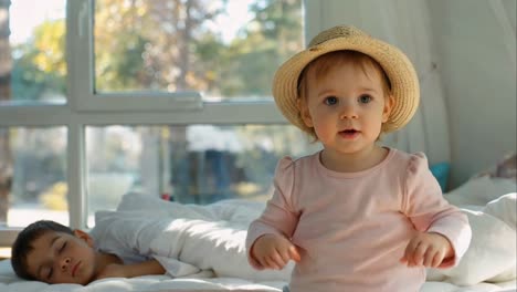 awake baby girl in hat beside sleeping boy in bed