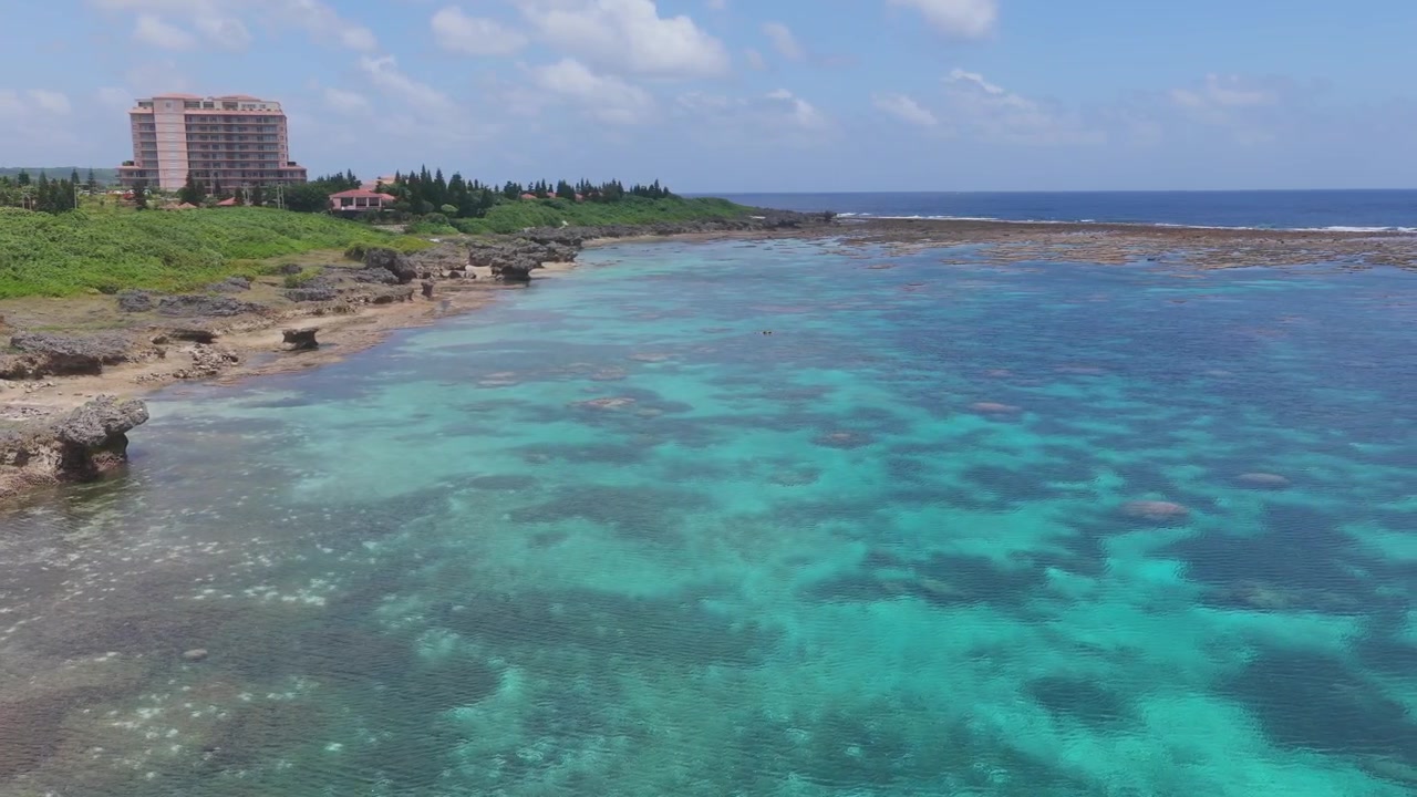 Inquadratura orizzontale, carrellata a destra da un'angolazione obliqua elevata, su acque turchesi e la costa rocciosa di Waiwai Beach