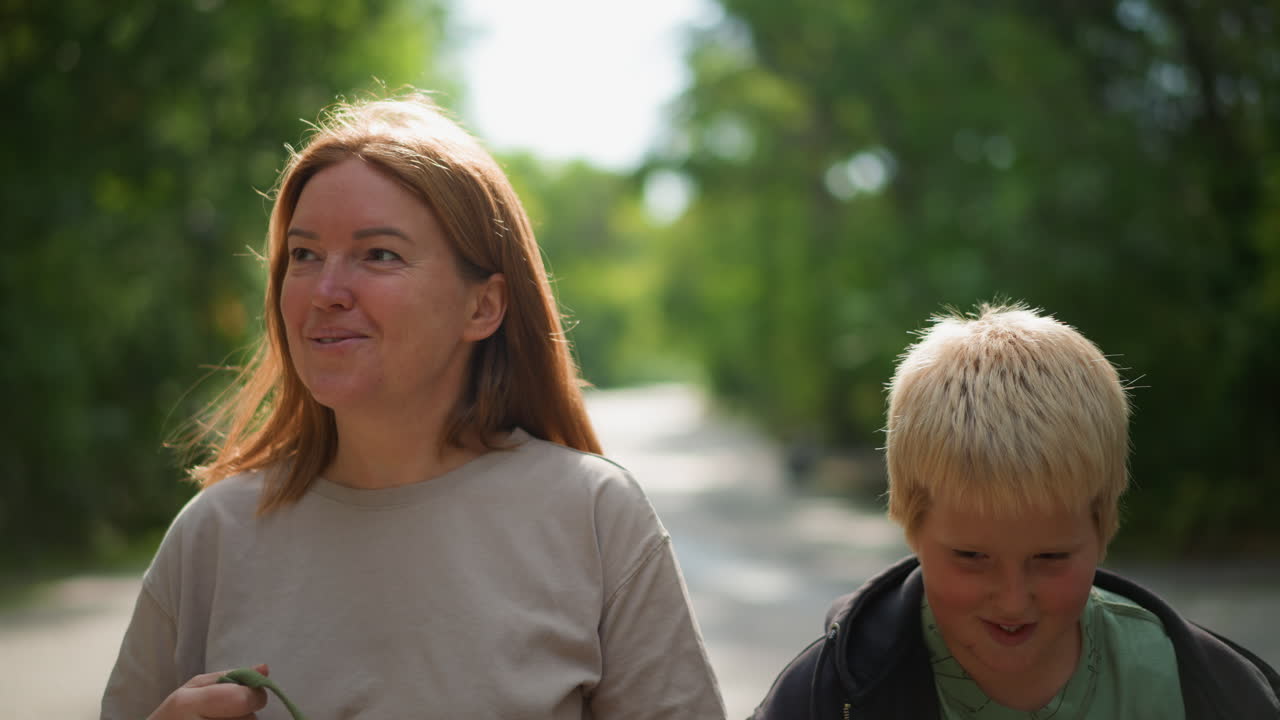 White Woman And Boy Walking Road, Mother Pointing Toward Treetops, Son Peeking Beside Her, Sunlight Filtering Through Green Canopy, Casual Conversation And Laughter, Warm Summer Mood, Candid Portrait