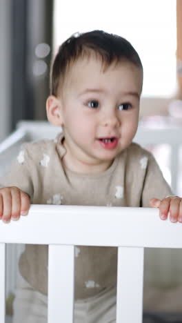 adorable caucasian kid stands inside his crib. beautiful child stretches hands in front of him demanding something. blurred backdrop. vertical video