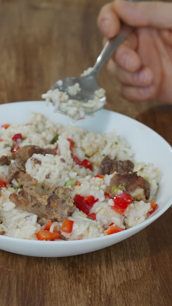 Man eats rice with meat and fresh vegetables using spoon. Male person sits at wooden kitchen table enjoying delicious meal for breakfast closeup