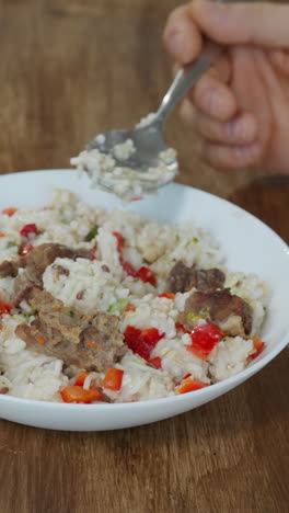 man eats rice with meat and fresh vegetables using spoon. male person sits at wooden kitchen table enjoying delicious meal for breakfast closeup