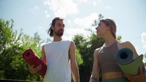 a blonde girl and a brunette guy in a summer sports uniform are walking through the park and getting ready for yoga in nature. the guy and the girl are specially mats for yoga and communicate in the park