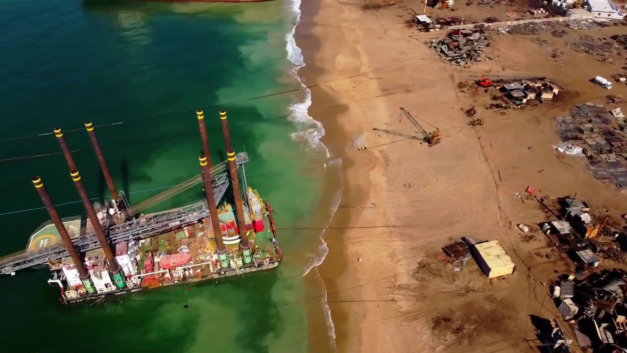 Drone video reveals massive piles of scrap metal and dismantled ships at Gadani ship breaking yard, Karachi. Daylight aerial footage highlights ocean pollution and human waste on the sandy coast.