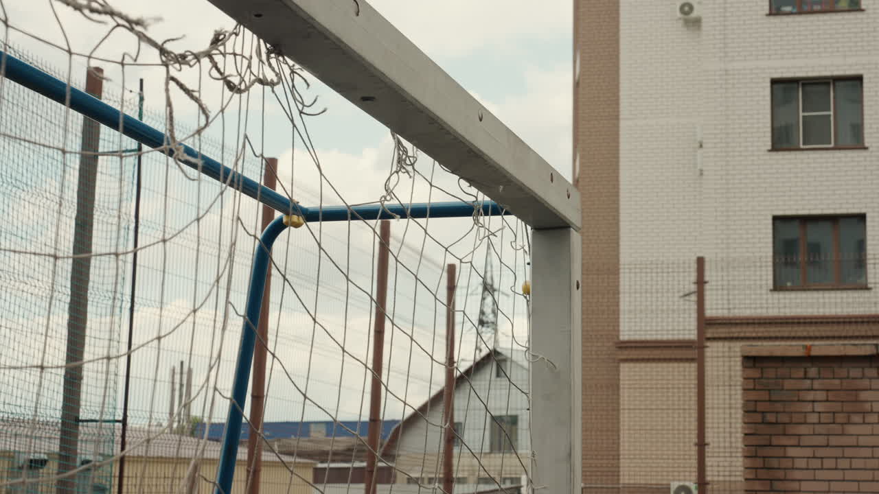 rain soaked football goal urban courtyard, metal frame and sagging net dotted with raindrops, weathered fence and apartment buildings in background, overcast sky casting muted light, puddles